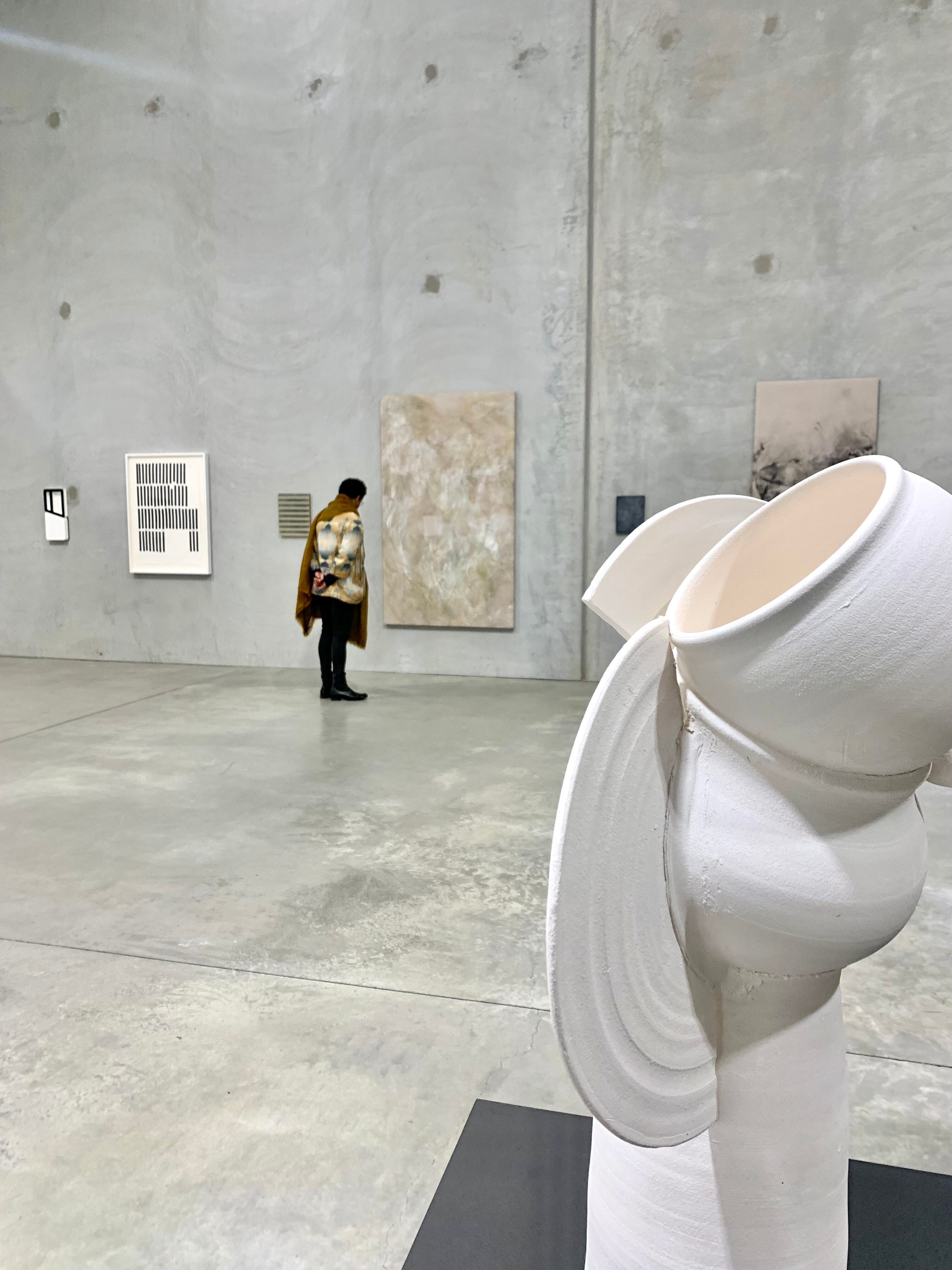 Round cream stone sculpture in foreground with artist in background observing large paintings on concrete slab wall