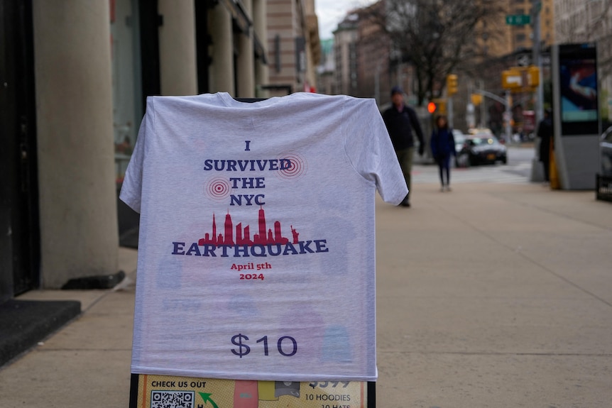 A t-shirt that says 'I survived the NYC earthquake April 5th 2024 $10' is hung over a a-frame sign on a NYC street
