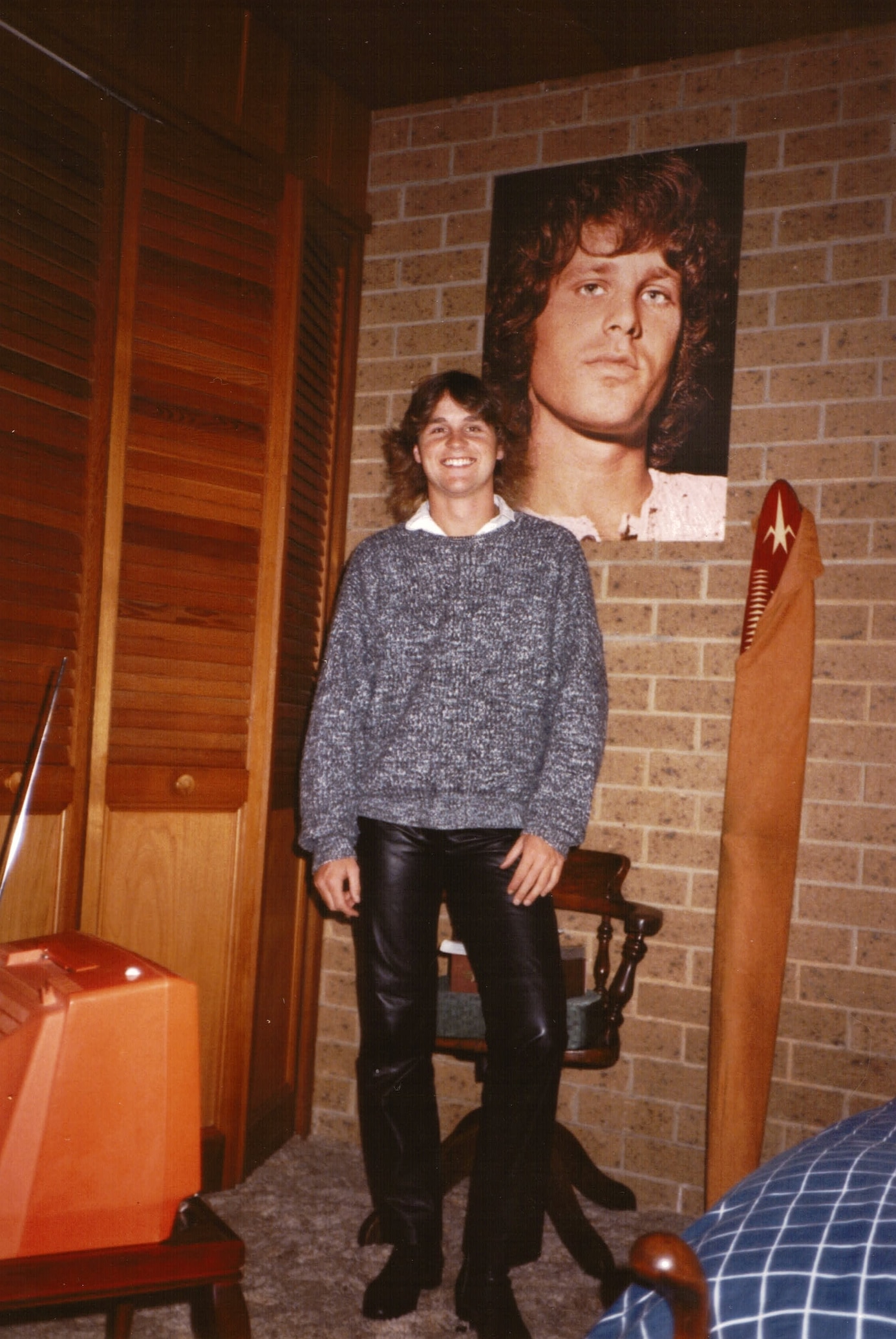 A young man wearing black pants and a blue jumper stands smiling beneath a poster of Jim Morrison.