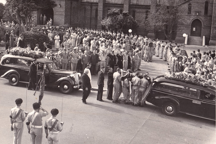 A old photograph of a group group of men holding up a casket near a car with crowds surrounding them.