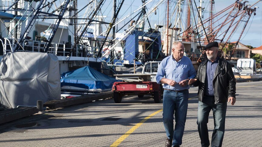 Two older men stroll in front of large fishing boats, deep in discussion