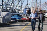 Two older men stroll in front of large fishing boats, deep in discussion