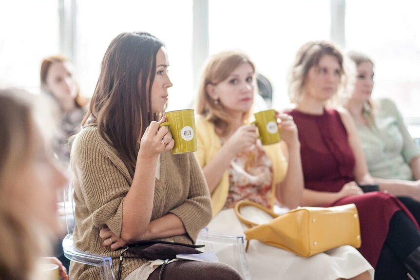 Women sitting in a meeting holding mugs and looking annoyed