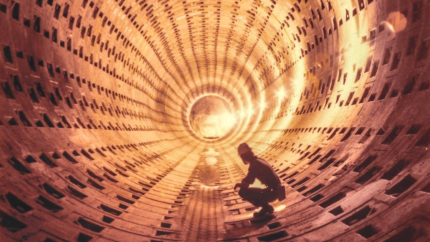 A man wearing a construction helmet squats in a large round concrete tunnel