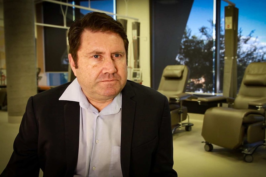 A man in a black suit jacket and shirt looks at the camera.