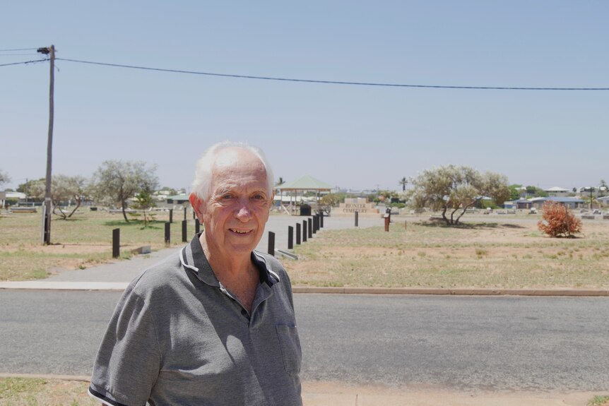 A man in his seventies with white hair stands across the road from a cemetery in the desert.