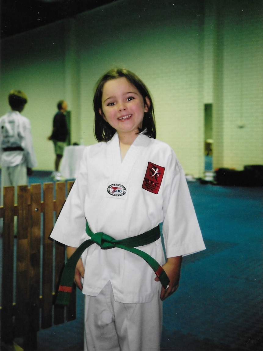 A supplied photo of Grace Scholl wearing a white karate uniform in a class, beaming at the camera.