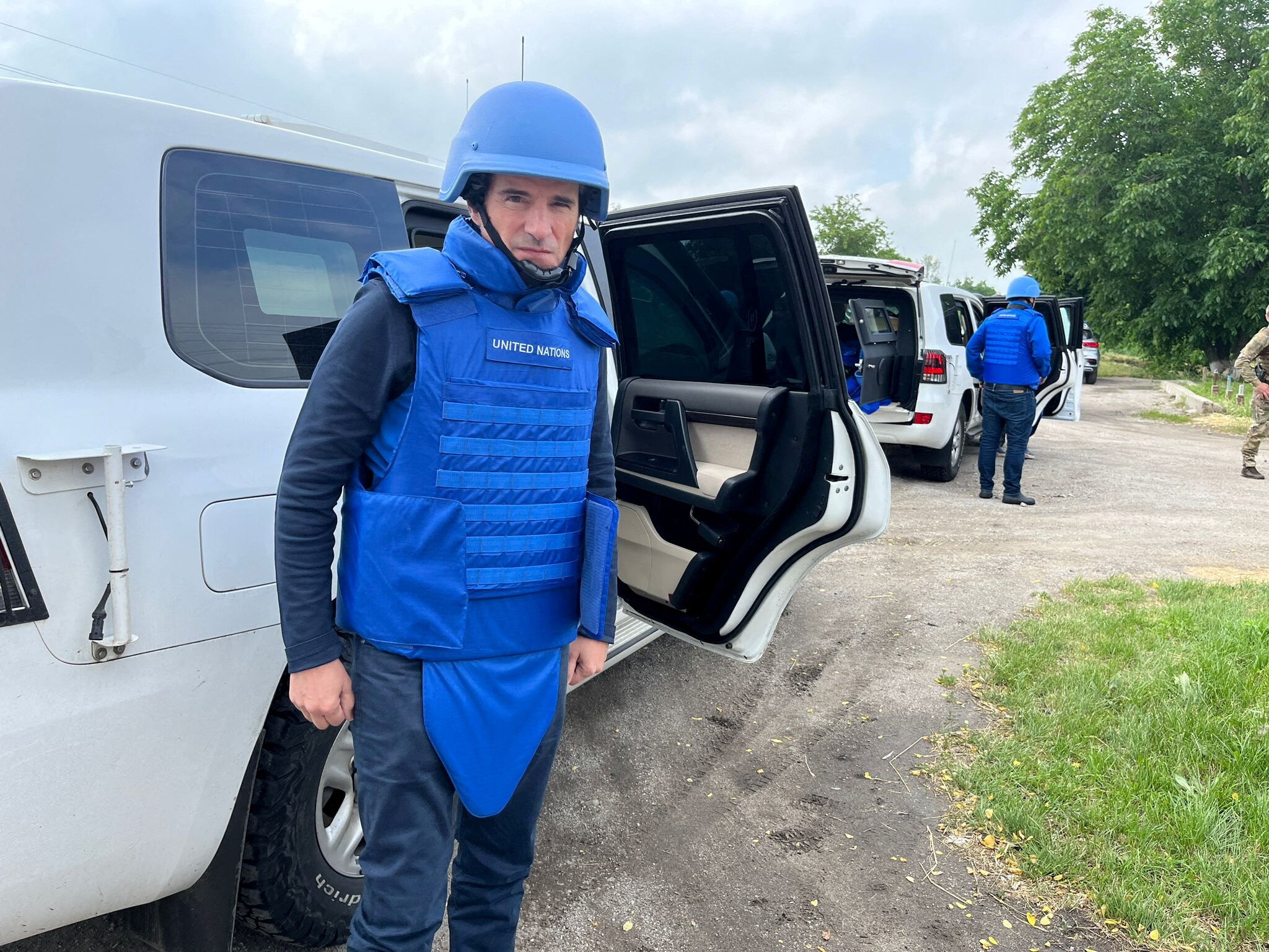 A middle-aged man in a bright blue helmet and bullet-proof vest stands next to a white armoured car.