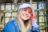 A young blonde woman grins while holding the brim of her baseball cap