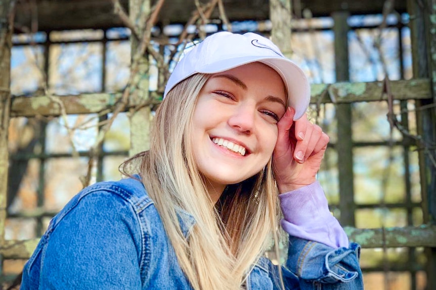 A young blonde woman grins while holding the brim of her baseball cap