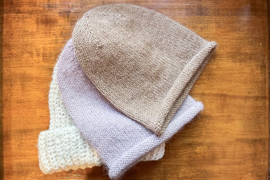 Three beanies (one cream, another lilac and a third tan) are seen laying atop each other on a wooden table.