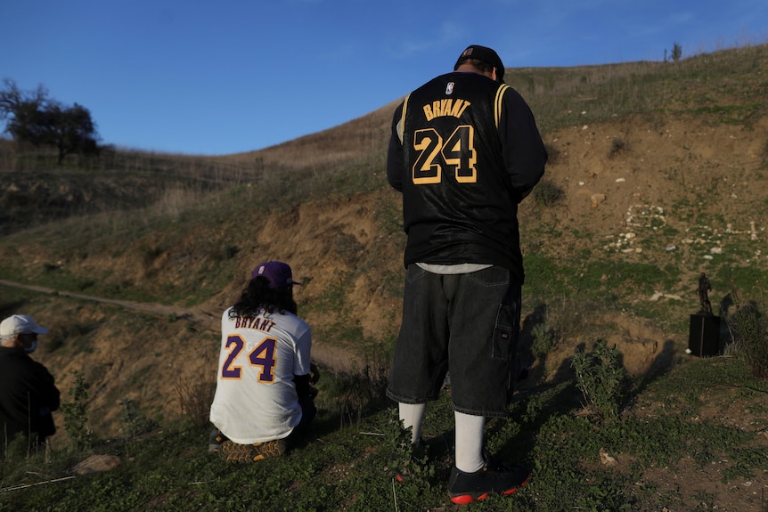 Two people wearing LA Lakers basketball jerseys look solemnly at a bronze statue on a hillside.