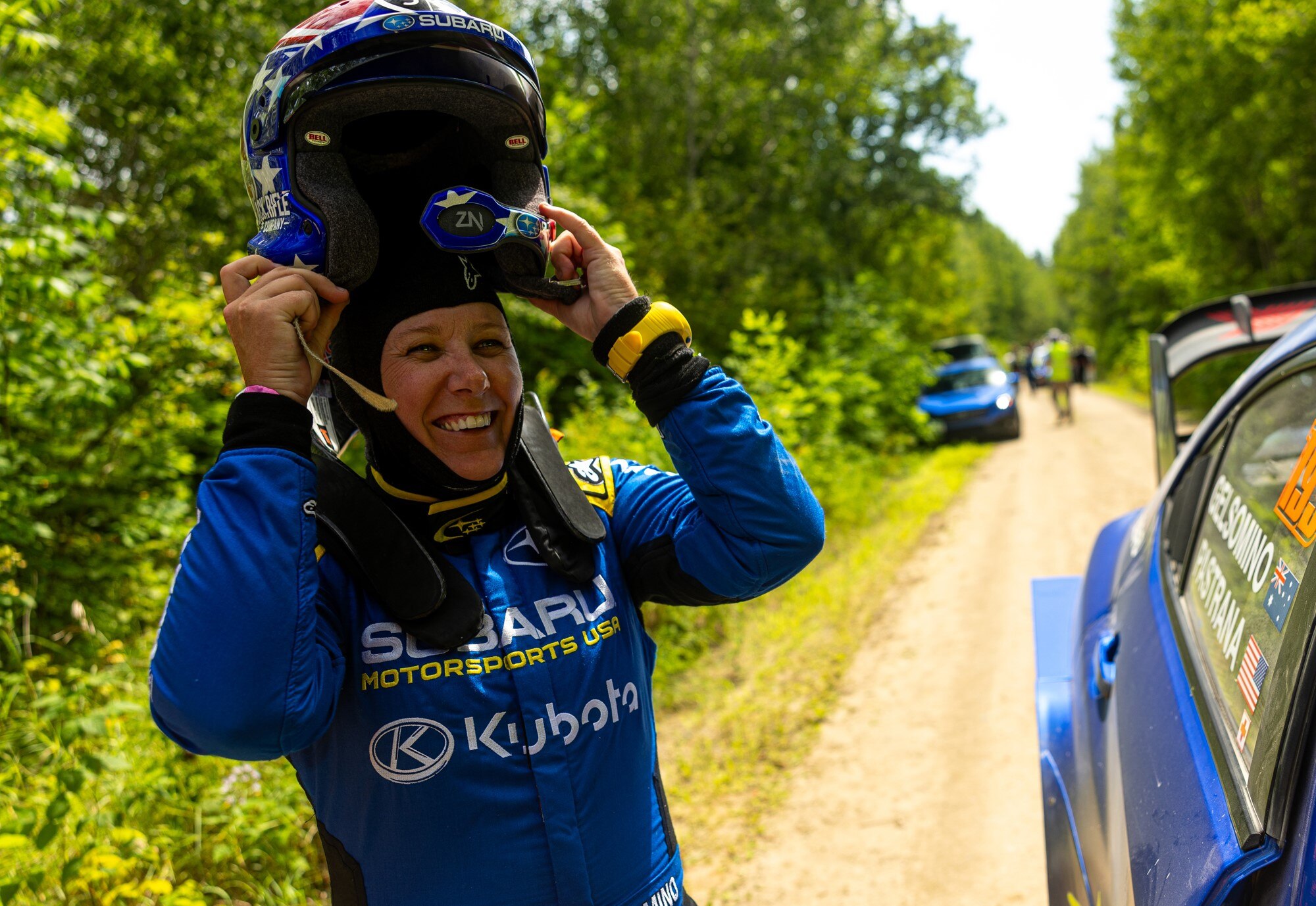 Žena koja nosi trkačku opremu i uklanja kacigu sa strane zemljane ceste s rally automobilima u pozadini.