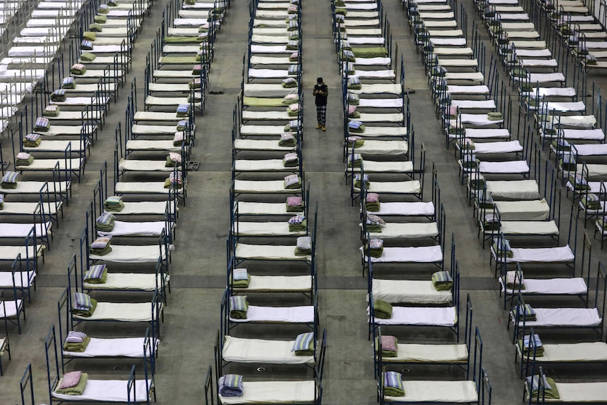 Un homme se tient au milieu de rangées de lits dans un grand centre de congrès.