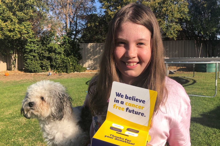 10-year-old Claire Anschuetz standing in her backyard with her fundraising box and her dog