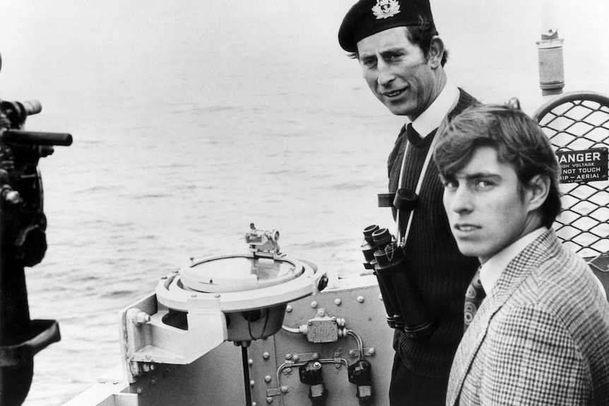 Prince Charles wearing a cap and Prince Andrew onboard a ship.