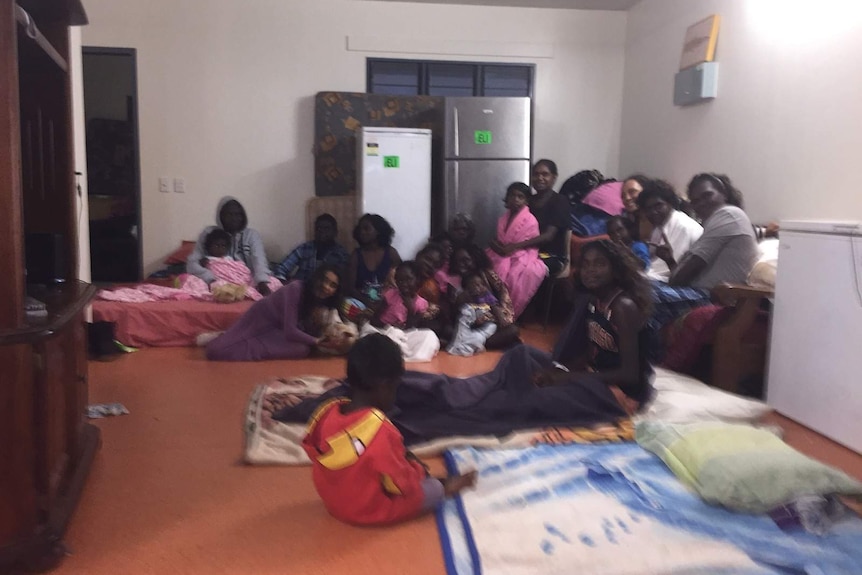 A group of mothers and daughters smile for a camera while sitting on a living room floor.