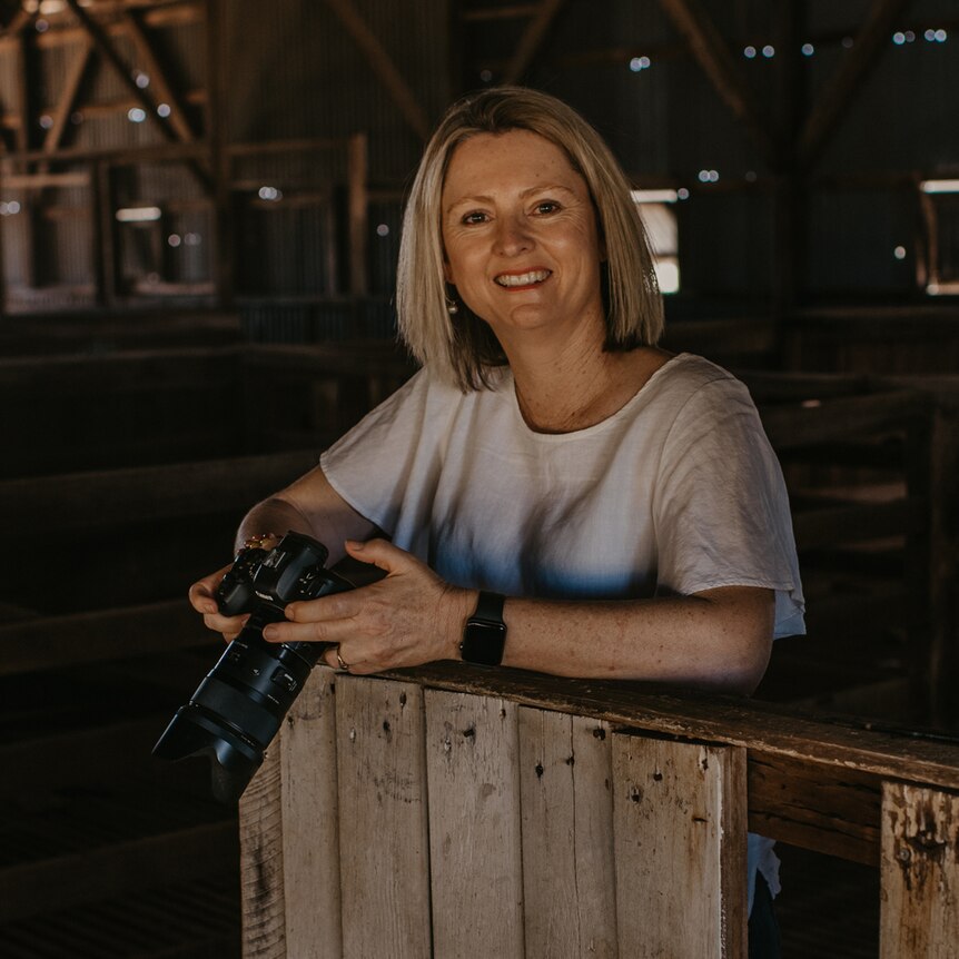 A blonde woman smiles for the camera, inside a shearing shed.