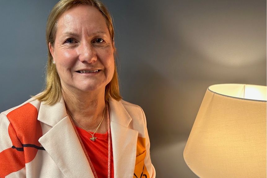 Blonde-haired woman in an orange and white jacket stands in a room smiling at camera.