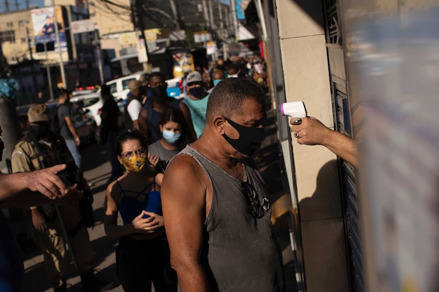 Man stands outside a building wearing a singlet and black face mask getting temperature checked. People are lined up behind him
