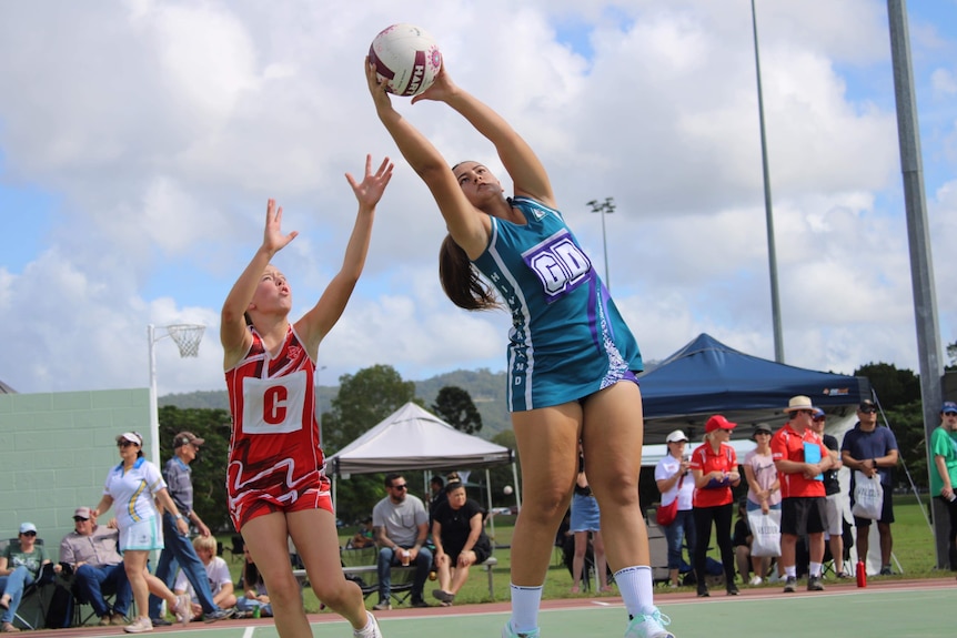 A girl in a blue netball uniform jumps with the ball to to throw it over the net, while another in red uniform tries to grab it.