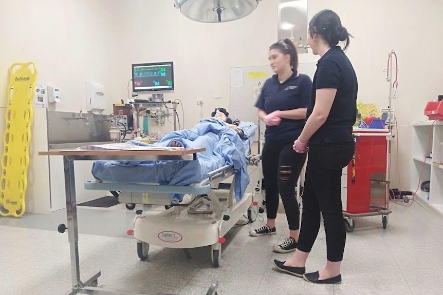 Two nursing students in black clothing stand in a hospital room next to a dummy patient propped on a hospital bed.