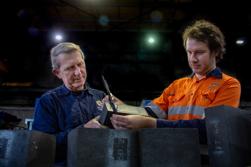 Two men wearing work shirts hold a dark block of material