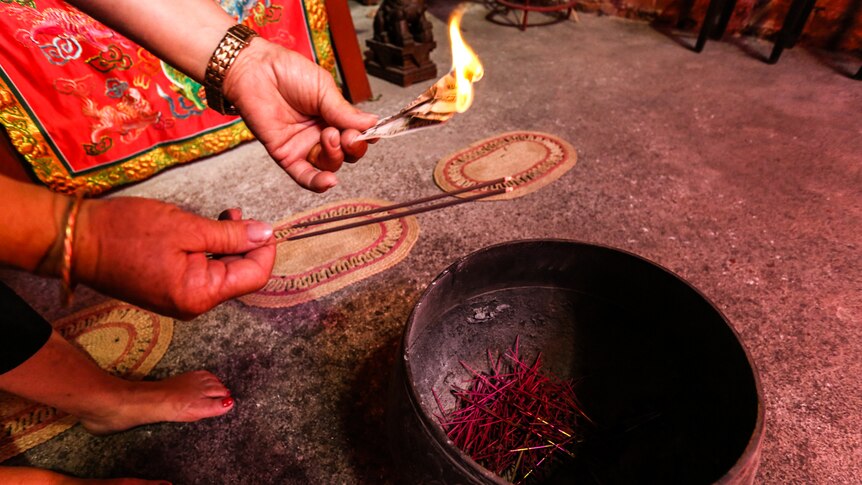 A woman's hands holding burning hell notes and holding burning incense over a brass bowl.