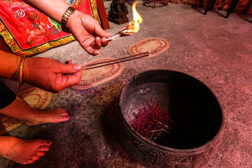 A woman's hands holding burning hell notes and holding burning incense over a brass bowl.