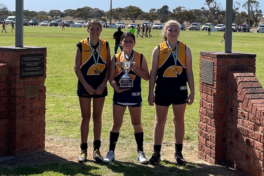 Three girls holding a grand final cup at a football ground in regional south australia.