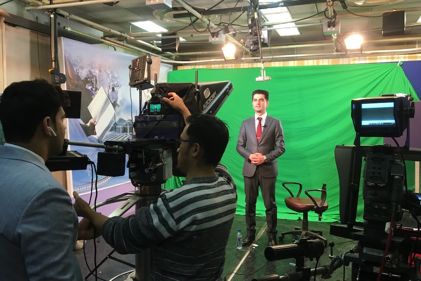 Khalid Amiri stands in front of a green screen while he presents the news for Radio Television Afghanistan.