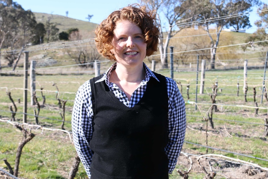 A woman stands in front of a vineyard in the sun.