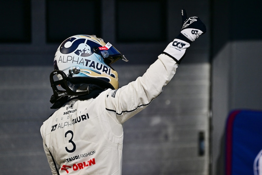 An F1 racing driver in full racing suit, waving to fans after hopping out of his car.