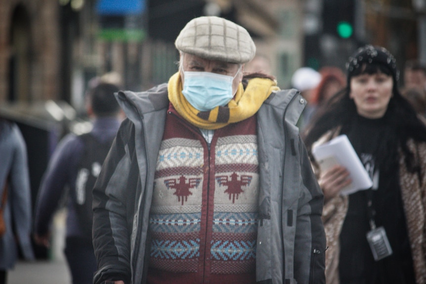 A man in a mask and warm clothing walks down a busy CBD street.