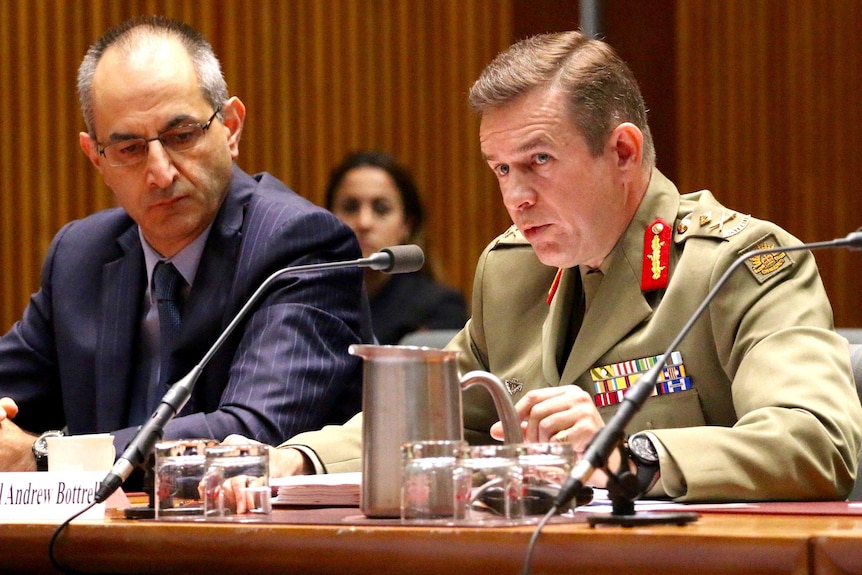 Major-General Andrew Bottrell, in uniform, sits behind a desk speaking into microphones.