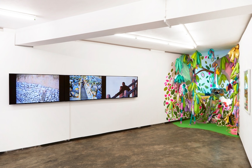 A three panel video work and a colourful installation sit next to one another in a white gallery space.