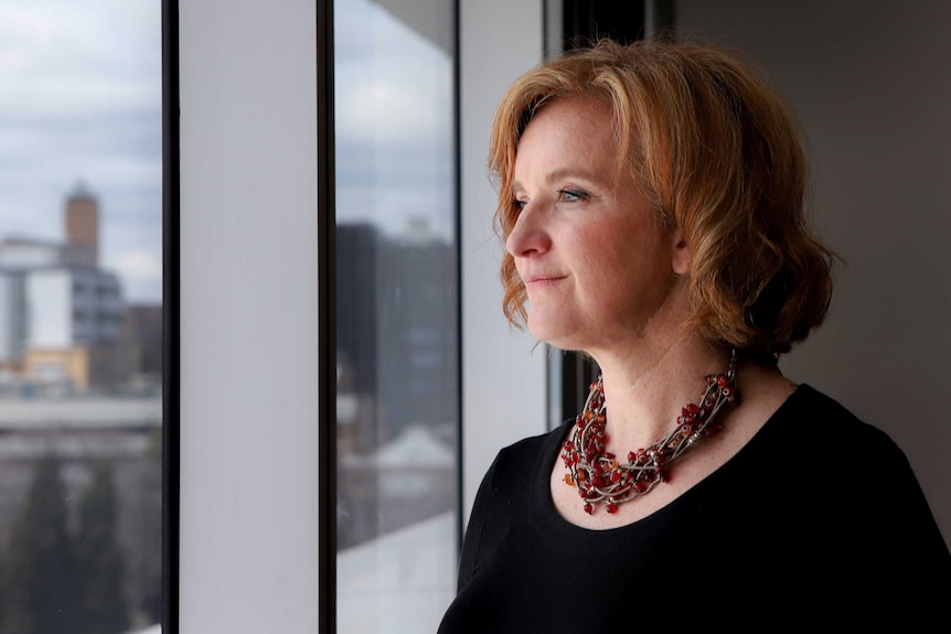 Woman with orange-red hair, black top and necklace stairs out window with city skyline in background