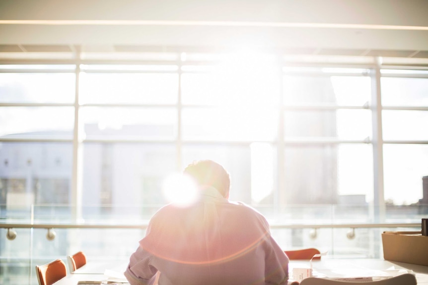 Man sitting in empty sunlit meeting room, depicting the stress and isolation that mismanaged workplace mental health can create.