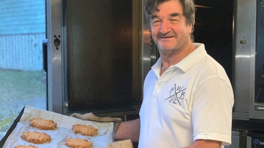 An image of a baker standing near an oven holding a tray of Anzac biscuits