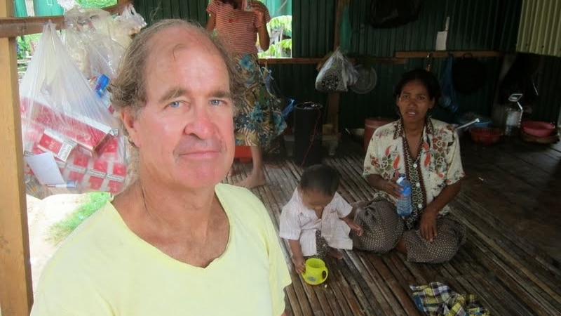 James Ricketson sits in a house in Cambodia with a woman and children in the background.