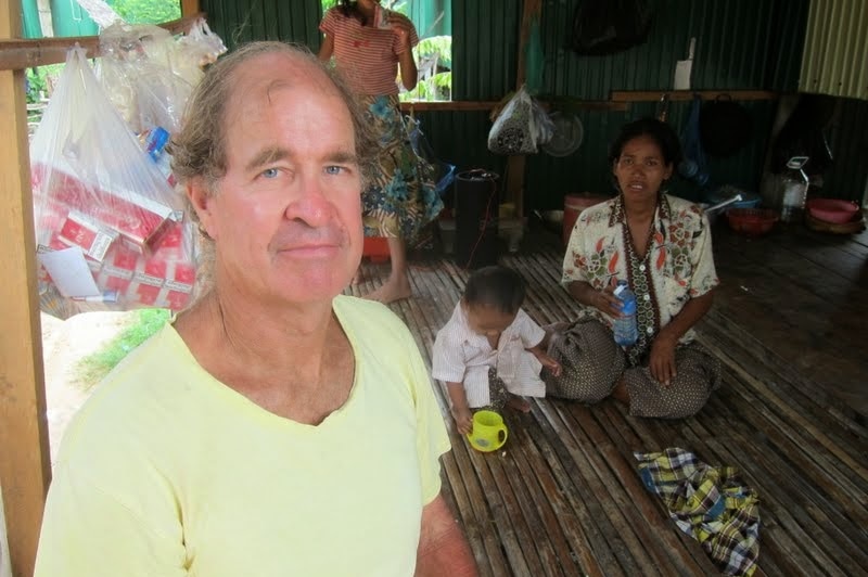 James Ricketson sits in a house in Cambodia with a woman and children in the background.