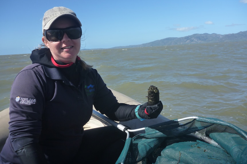 A woman in warm jacket, sunglasses, hat and gloves sits on a boat in the ocean, holding a large dugong poo in her hand