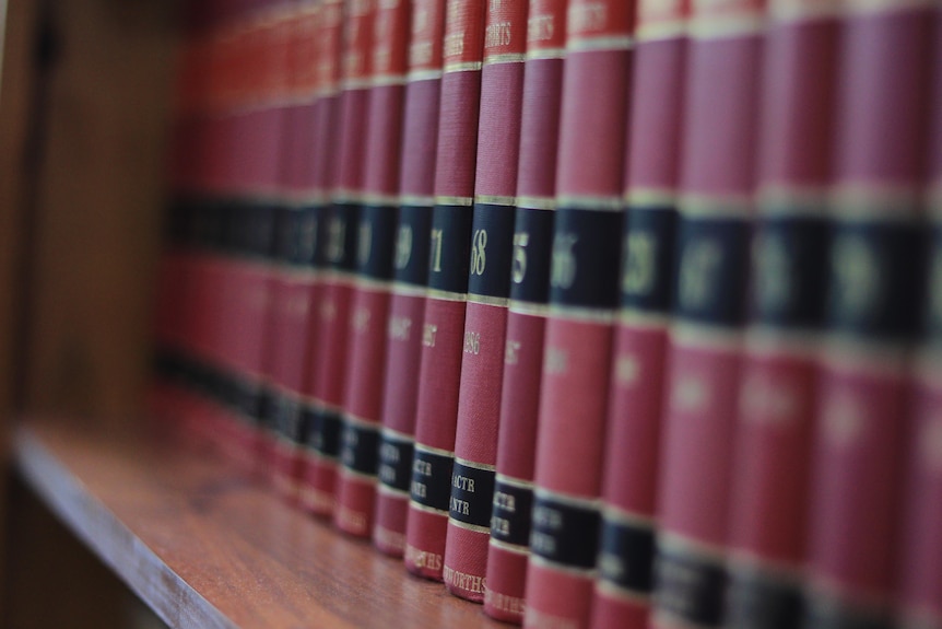 Red books sit lined on a wooden shelf