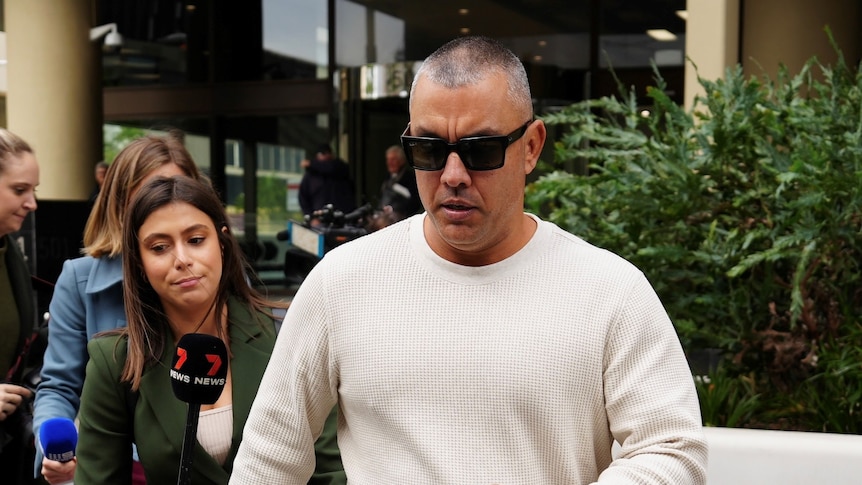 A man in a white jumper walks out of court with black sunglasses