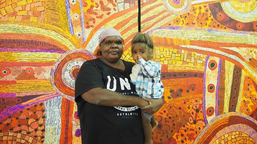 An Aboriginal woman and a small child stand in front of a large dot painting.