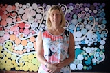 A middle aged woman stands in front of a colourful dot artwork with her hands together, wearing a dress.