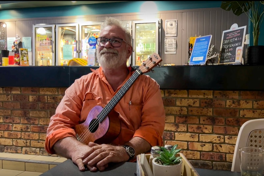 Kent sits in front of the camera smiling, holding his ukulele in his lap with the neck pointing up.