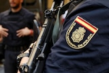 A close-up of a Spanish police officer's shoulder badge and gun with another officer and civilians in the background.