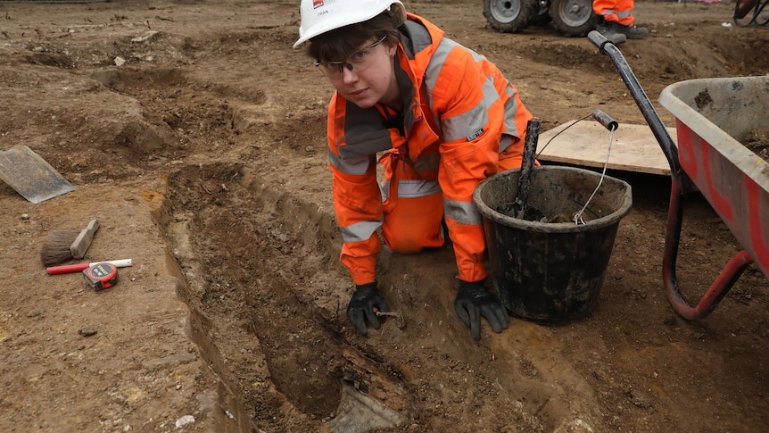 A woman in a hardhat, safety glasses and high-vis clothes leans over a long, narrow hole in the dirt, next to a bucket