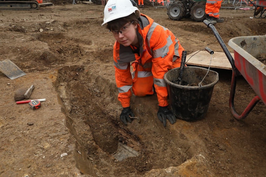 A woman in a hardhat, safety glasses and high-vis clothes leans over a long, narrow hole in the dirt, next to a bucket